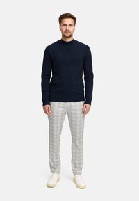 Mann steht mit dem Gesicht nach vorne, trägt einen dunkelblauen Pullover, hellgraue karierte Hose und cremefarbene Sneakers vor einem einfarbigen Hintergrund.