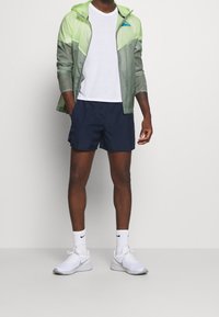 Letvægts, grøn og grå vindjakke med lynlås; hvid T-shirt; marineblå shorts; hvide sneakers med grå såler; atletiske sokker.