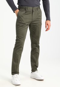 Pantaloni verde oliva realizzati in tessuto liscio, con una vestibilità sartoriale, tasche anteriori e una vita classica. Abbinati a sneakers bianche.