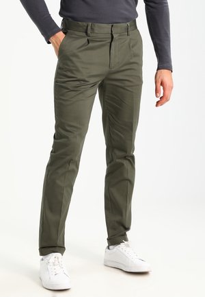 Pantaloni verde oliva realizzati in tessuto liscio, con una vestibilità sartoriale, tasche anteriori e una vita classica. Abbinati a sneakers bianche.