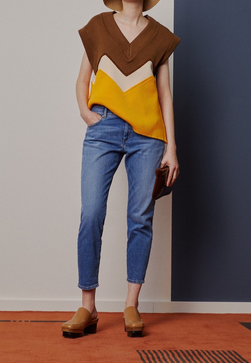 Hnědý, žlutý a krémový pletený svetr s výstřihem do V a vzorem ve tvaru šipek. Světle modré džíny a hnědé dřeváky. Drží hnědou peněženku.
