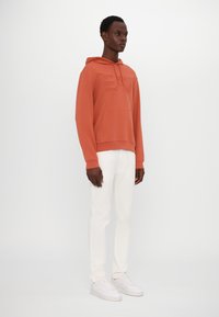 Felpa con cappuccio color terracotta con logo testurizzato, tasca frontale, abbinata a pantaloni bianchi e sneakers bianche, su uno sfondo neutro.
