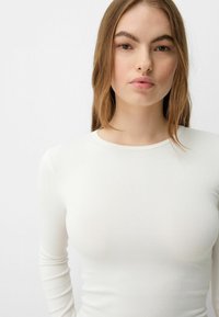 Bela dolga majica s dolgimi rokavi, prilegajočega kroja, rebraste teksture, okroglo izrezana, elastična tkanina, podrobno šivanje. Izgleda elegantno in prilegajoče.