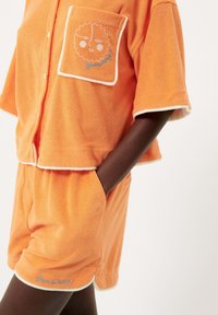 Oranje badstof set bestaande uit een shirt met korte mouwen en een zak, voorzien van een geborduurd gezichtsontwerp, en shorts met zijzakken.