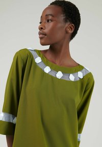 Groene tuniek met een ronde halslijn, voorzien van een doorschijnende grijs-witte geometrische accent langs de halslijn en mouwen, met een zachte textuur.