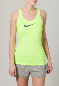 Neonowo zielony top Nike na ramiączkach z okrągłym dekoltem i czarnym logo Swoosh; w zestawie z szarymi sportowymi szortami. Gładka faktura i dopasowany krój.