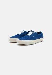 Blue suede sneakers with white laces, low-profile design, and a cream rubber sole. Logo tag visible on the side, smooth texture.