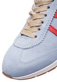 Gros plan sur une sneaker en daim bleu clair avec des lacets tressés beige et un détail cousu rouge sur le côté au-dessus de la semelle blanche.