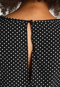 Mustaa kangasta, jossa on valkoisia pilkkuja, takaosassa avaus, joka on kiinnitetty mustalla napilla. Sujuva rakenne.