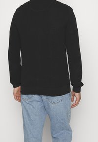 Man som bär en strukturerad svart tröja och ljusblå jeans, står med ryggen mot kameran mot en enfärgad bakgrund.