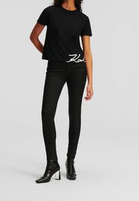 Czarny T-shirt z krótkim rękawem z białym logo "Karl" na dolnym brzegu, w zestawie z czarnymi legginsami w wzór i czarnymi botkami za kostkę z metalicznymi akcentami na obcasach.