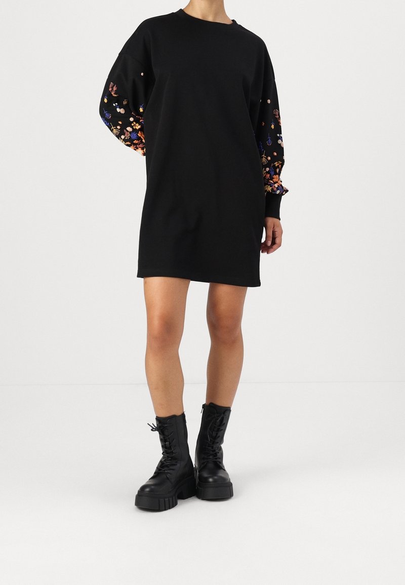 Robe sweat-shirt noire avec manches à broderies florales et poignets côtelés, portée avec des bottes plateformes noires à lacets. Texture lisse sur toute la surface.