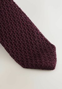 Cravatta lavorata a maglia di colore viola scuro con un motivo strutturato e intrecciato. La punta è sottile e leggermente appuntita, con una finitura lucida e ricca.