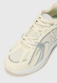 Light cream athletic shoe with mesh fabric and silver accents. Features rounded toe, white laces, and supportive sole. Textured detailing visible.