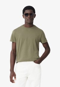 T-shirt in cotone verde oliva con collo rotondo, caratterizzata da un piccolo logo ricamato sul petto. Abbinata a pantaloni bianchi e occhiali da sole scuri.