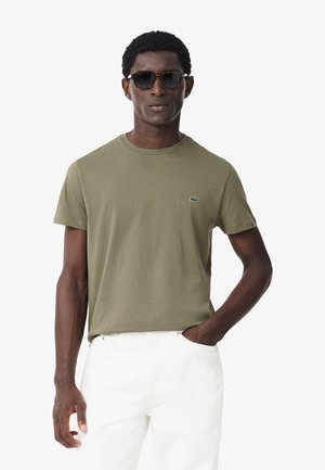 T-shirt in cotone verde oliva con collo rotondo, caratterizzata da un piccolo logo ricamato sul petto. Abbinata a pantaloni bianchi e occhiali da sole scuri.