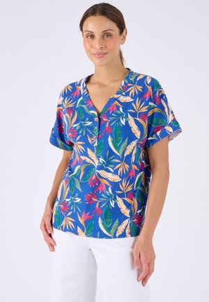 Donna che indossa una camicetta blu a maniche corte con stampa multicolore di foglie tropicali e pantaloni bianchi, in piedi davanti a uno sfondo semplice.