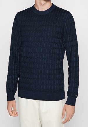 Mann trägt dunkelblauen, strukturierten Zopfpullover und weiße Hose, steht mit locker hängenden Armen an den Seiten vor schlichtem Hintergrund.