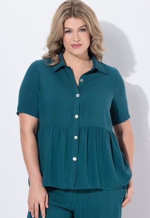 Kobieta o blond włosach do ramion, ubrana w turkusową koszulę na guziki z krótkim rękawem i dopasowane spodnie, stoi na tle białego tła.