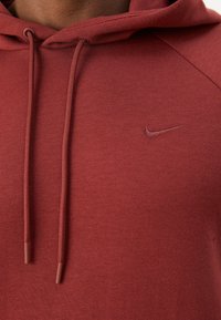Detailní záběr na vínovou mikinu Nike s kapucí a šňůrkami, s vyšívaným logem swoosh na hrudi.