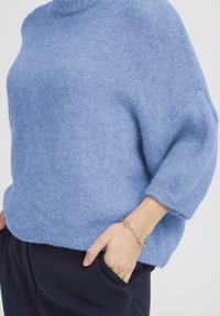 Pull en tricot bleu clair avec un col montant, des épaules tombantes et une coupe décontractée. La texture est duveteuse, assortie à un pantalon foncé et un bracelet.