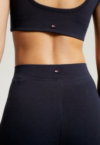 Czarny stanik sportowy i legginsy z wysokim stanem o gładkiej teksturze. Posiada małą metkę z logo z czerwonymi, białymi i niebieskimi akcentami na pasie.