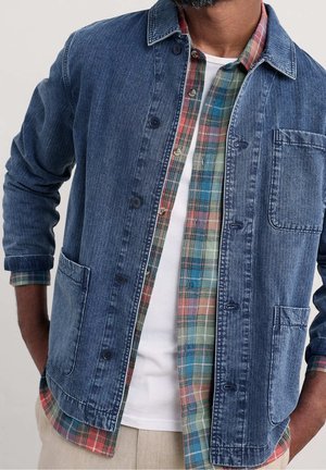 Mand iført en blå denimjakke over en flerfarvet ternet skjorte og en hvid t-shirt, hænderne i lommerne, vist fra nakke til talje.