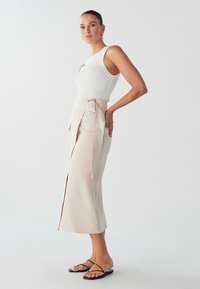Bela rebrasta majica bez rukava u kombinaciji s pamučnom midi suknjom boje bež s visokim strukom, koja ima remen i prednji prorez, te crnim ravnim sandalama.