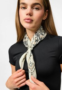 Lichte crèmekleurige sjaal met zwart en beige bloemenpatronen, om de nek geknoopt, tegen een zwarte top; zachte textuur en gebogen vorm.