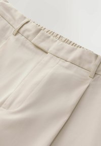 Ceinture de pantalon beige avec dos élastique, passants pour ceinture et détail de poche avant sur tissu lisse.