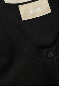 Svart kofta med V-hals och en enda svart knapp. Har en mjuk textur och en beige etikett med "JJXX" i vit stil.