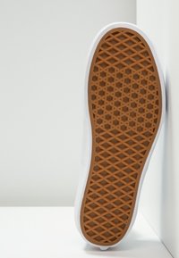 Semelle d'une chaussure blanche avec un motif de grip en forme de gaufre marron, positionnée à la verticale contre un mur et une surface blancs.