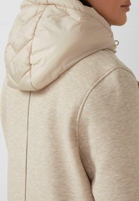 Beige sweatshirt met een zachte textuur, met een lichte, gewatteerde capuchon. Het ontwerp heeft een nette naad aan de achterkant en ronde schouders.