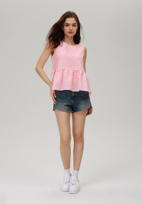 Top peplum in gingham rosa senza maniche, abbinato a pantaloni corti in denim blue. La modella indossa sneakers bianche e calze corte. Sfondo semplice.