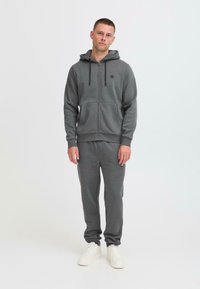 Haut à capuche gris zippé avec des cordons et un logo, associé à un pantalon de survêtement assorti. Le matériau semble doux avec une coupe décontractée et des poignets côtelés.