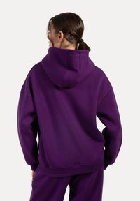 Sudadera con capucha púrpura hecha de un tejido suave. Presenta una capucha espaciosa, hombros caídos y puños acanalados. Textura suave con un detalle mínimo.