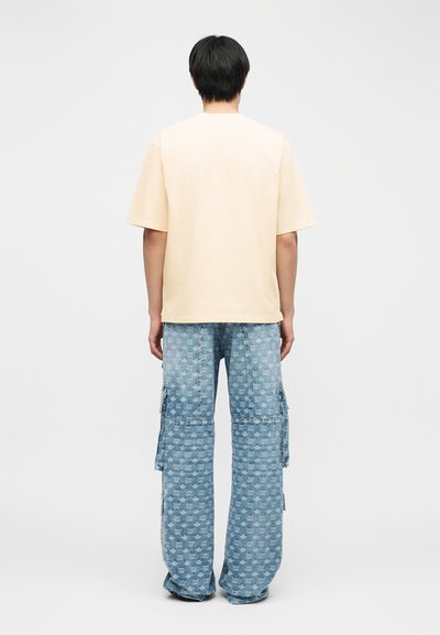 Άνδρας που φοράει μονόχρωμο μπεζ πουκάμισο με κοντά μανίκια και μπλε παντελόνι cargo με φαρδιά πόδια και σχέδιο, στέκεται γυρισμένος με την πλάτη προς το λευκό φόντο.