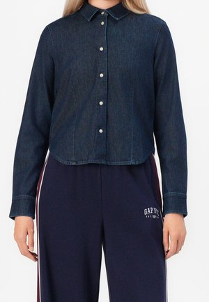 Kvinde iført en mørkeblå denim skjorte med knapper og lange ærmer samt marineblå GAP NYC bukser med hvide og røde sidelister.
