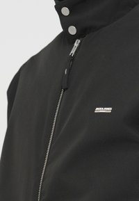 Μαύρο μπουφάν με ψηλό γιακά, ασημένιο φερμουάρ και κουμπιά τύπου σναπ. Διαθέτει μικρό μεταλλικό λογότυπο στο αριστερό στήθος. Λεία υφή υφάσματος.