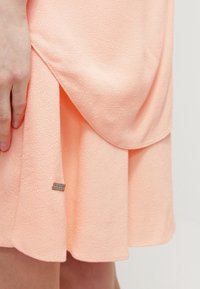 Pfirsichfarbenes Kleid aus strukturiertem Stoff, mit einem mehrlagigen Saum und einem kleinen metallischen Logoakzent in der Nähe des unteren Saums.