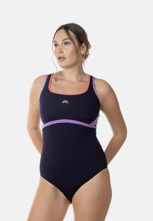Donna con un costume da bagno nero con rifiniture rosa e viola, che guarda di lato con le mani posizionate dietro la schiena.