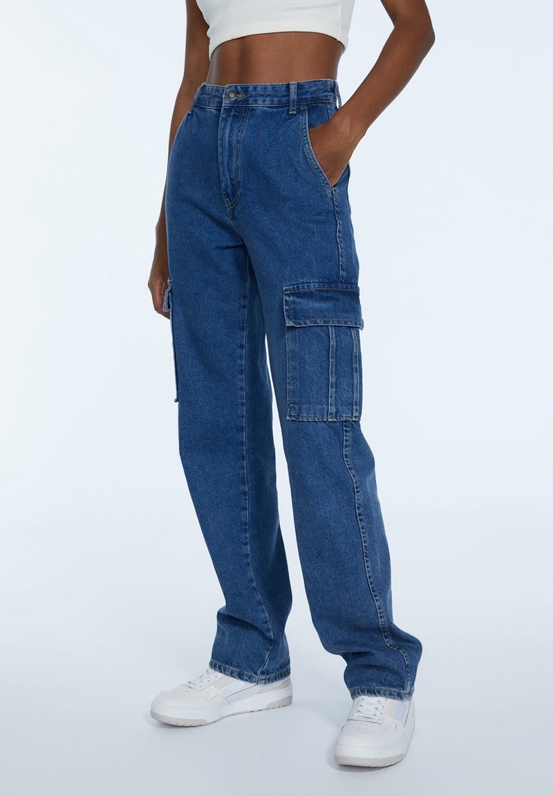Personne portant un pantalon cargo en denim bleu avec de grandes poches latérales, un top court blanc et des baskets blanches, debout avec une main dans la poche.