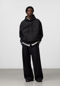 Felpa con cappuccio grigio scuro in pile con tasca frontale, abbinata a pantaloni neri larghi. Il tessuto appare morbido con una finitura testurizzata.