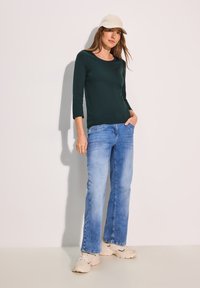 Tumma vihreä kolmonenesosa hiha toppi, vaaleansiniset farkut, beige lippalakki ja valkoiset lenkkarit. Pehmeä tuote, rento istuvuus ja epämuodollinen tyyli.