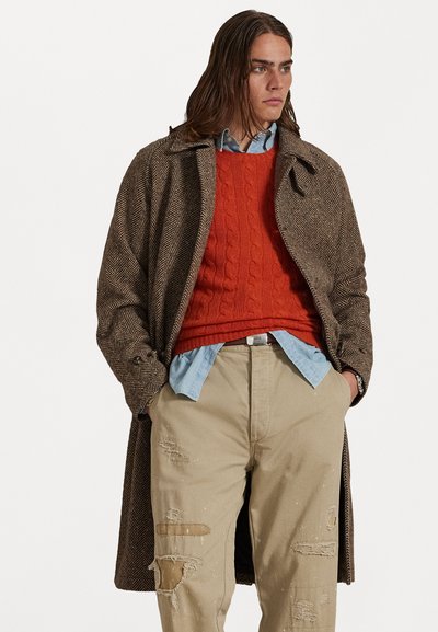 Manteau en chevron marron sur un pull rouge en tricot torsadé et une chemise bleu clair. Pantalons beiges avec des détails usés. Figure aux cheveux longs se tient droit.