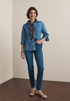 Femeie stând și zâmbind, purtând o cămașă din denim albastru, blugi skinny, eșarfă cu model la gât, brățară neagră și sandale negre cu degetele descoperite.