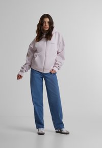 Felpa rosa chiaro con zip, tasca frontale e logo ricamato, abbinata a jeans azzurri a vestibilità rilassata e scarpe grigie.