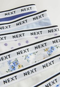 Pile de sous-vêtements pliés avec ceintures élastiques, chacun étiqueté "NEXT", en motifs unis, à pois et floraux sur des fonds clairs.