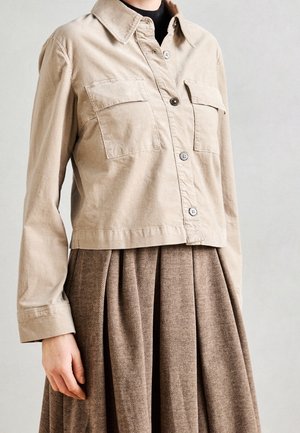 Personne portant une veste beige boutonnée avec des poches poitrine sur une jupe plissée marron à motif en chevrons.