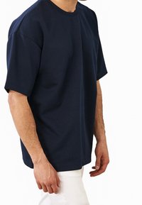 Hombre vestido con una camisa de manga corta azul marino de corte holgado y pantalones blancos, mostrado de los hombros a la mitad del muslo, contra un fondo blanco.